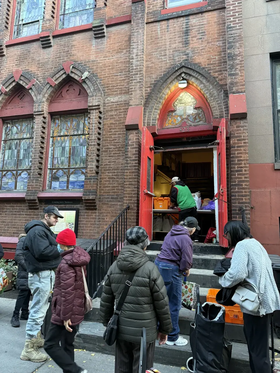 A pantry morning at St. Clement’s: the red doors open, volunteers at the tables inside, and neighbors waiting on the steps