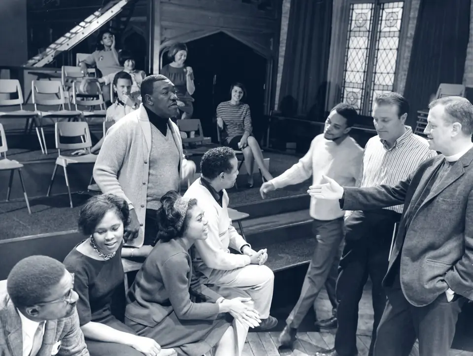 Actors gathered on the stage and risers at St. Clement’s in the 1960s, with a priest in a clerical collar among them