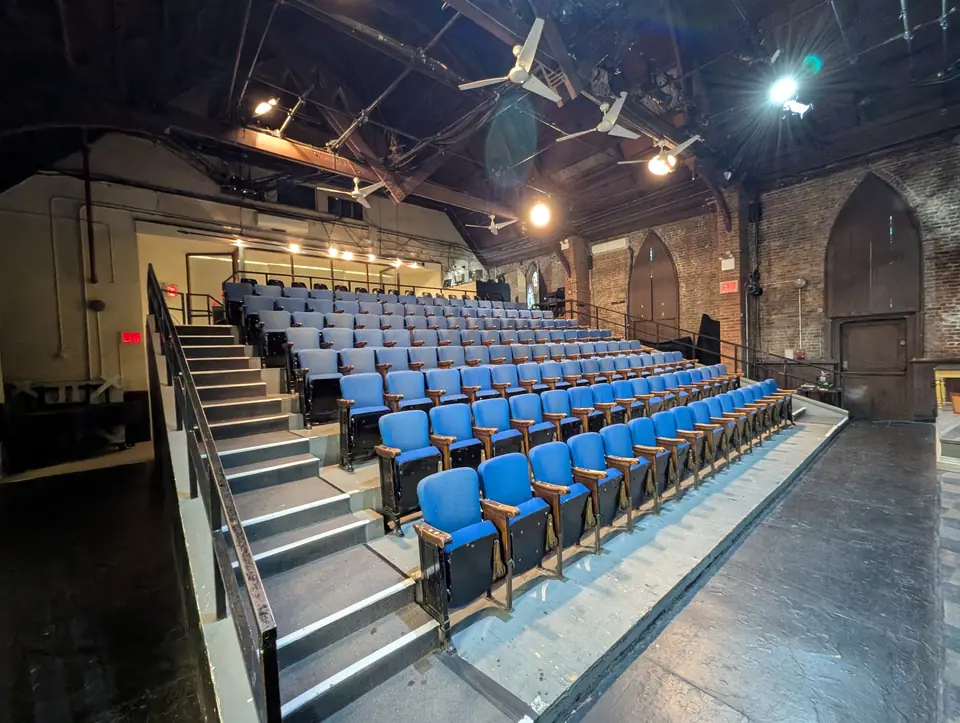 The theater’s blue seats rising on their risers beside the stairs, under the old church roof and its brick Gothic arches